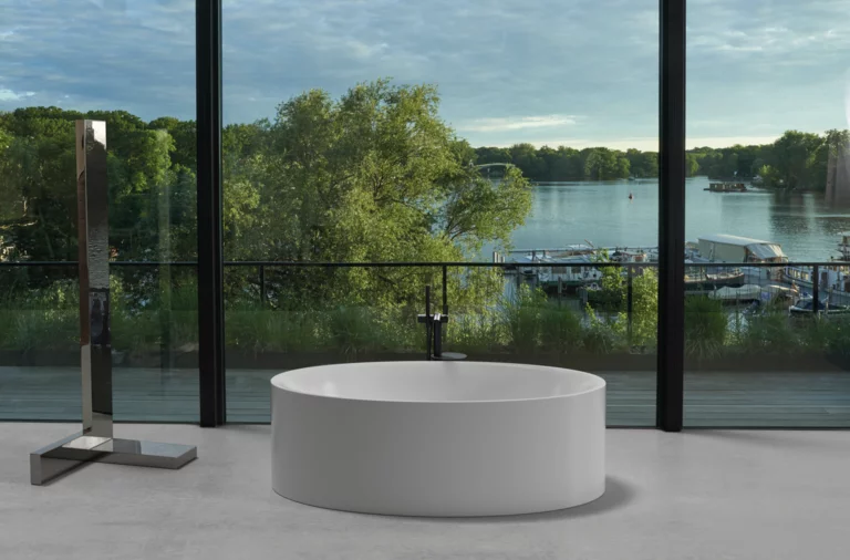 Freistehende, ovale Badewanne vor Panoramafenster mit Blick auf See, Steg und grüne Uferlandschaft.